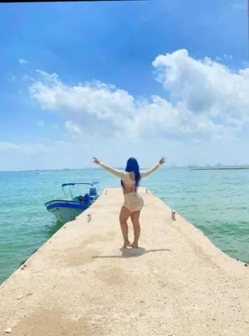 Woman in a white bikini standing on a pier with arms outstretched against a bright blue sky and ocean view.
