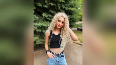 Smiling woman with curly blonde hair wearing a black tank top and denim jeans in a lush green outdoor setting.