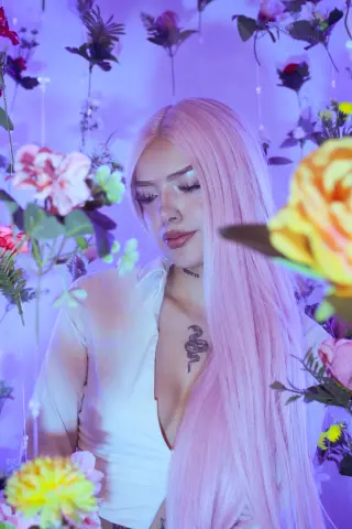 BabyVelvett with long pink hair poses in a white shirt surrounded by colorful flowers.