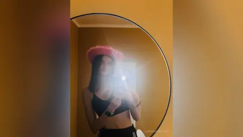 Young woman in a black crop top and pink fuzzy hat taking a mirror selfie in a warm, sunlit room.