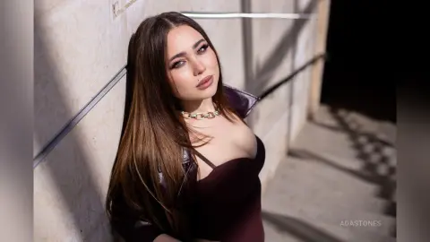 Glamorous woman in a dark burgundy outfit and silver chain necklace leaning against a sunlit stone wall.