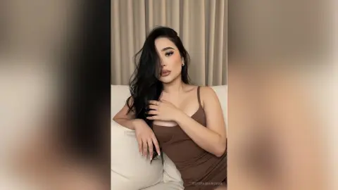 Young woman with long dark hair wearing a brown ribbed tank top sitting on a white sofa in a bright room.