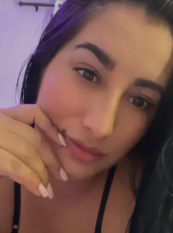 Close-up portrait of a young woman with long dark hair and manicured nails in soft indoor lighting.