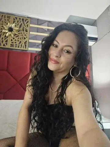 Smiling woman with long curly hair wearing a black top and gold hoop earrings in a modern bedroom.