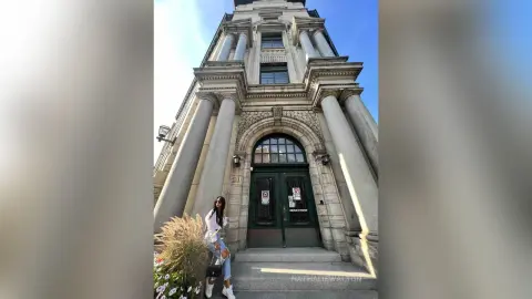 Stylish woman in a white shirt and jeans standing before a grand classical stone building entrance.