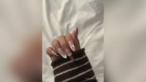 Close-up of elegant almond-shaped French tip manicure with gold nail art on hands wearing a cozy brown sweater.