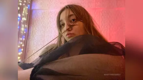 MartineKibbey poses with long brown hair, wearing a sheer black top that shows her breasts against a pink background with string lights.