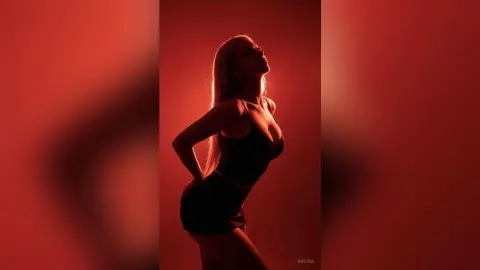 Kalira poses in profile against a red background, wearing a black tank top and shorts that highlight her curves.