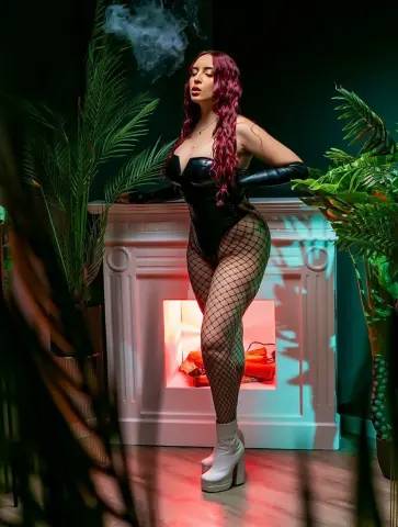 Red-haired woman in black bodysuit and fishnets posing by a white fireplace in a moody, tropical indoor setting.