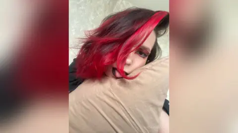 AndreaHazel poses with her bright red hair falling over her face while she bites a tan pillow.