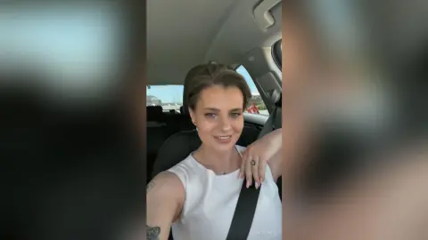 AnishaBee smiles while sitting in a car seat wearing a white tank top, with her hand resting near her neck.