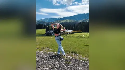 LesleyRed poses in a black crop top and light blue jeans against a scenic mountain landscape.