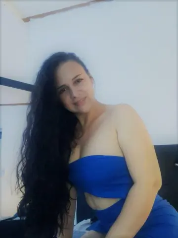 BrianaCharlot poses in a blue tube top and matching blue skirt, letting her long, dark wavy hair fall over one shoulder against a plain white wall.