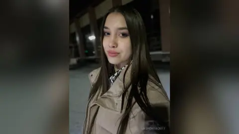 Young woman in a beige winter coat smiling outdoors at night in a moody urban setting.