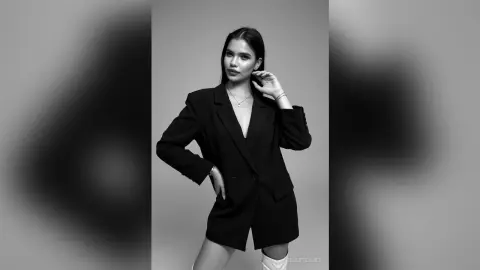 Elegant woman in a black blazer and necklace posing against a minimalist grey studio background.