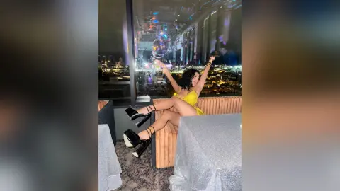 NicoleBella poses in a yellow dress and black platform heels, leaning back against a tan sofa in a room with a city view.