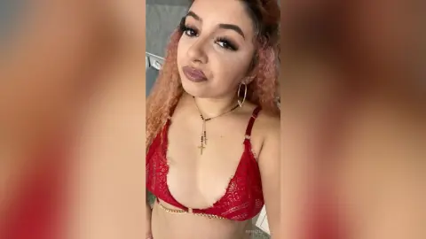 Young woman with wavy hair wearing a red lace bikini and gold cross necklace in a soft indoor setting.
