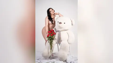 Nude woman with long dark hair posing next to a giant white teddy bear and red roses in a bright studio.