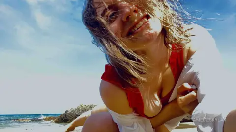 Smiling young woman in a red bikini and white cover-up relaxing on a sunny beach under a blue sky.