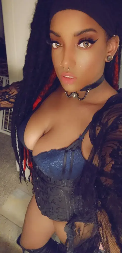 Shorty_Laveau poses with a direct gaze, wearing a black spiked choker and dark eye makeup against a dark background.