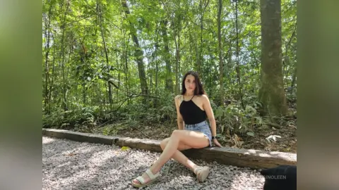 RossiNoleen sits on a fallen log in a lush green forest wearing a black halter top and denim shorts.