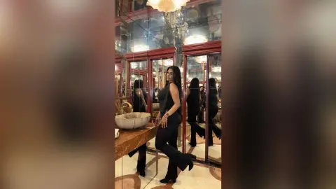 Elegant woman in a black sleeveless top and trousers posing in a luxurious mirrored room with warm lighting.