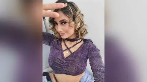 Glamorous woman in a purple sparkly crop top making a peace sign in a bright indoor setting.