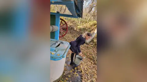 Woman in black outfit carrying a metal bucket through autumn leaves near a rustic blue wooden structure.