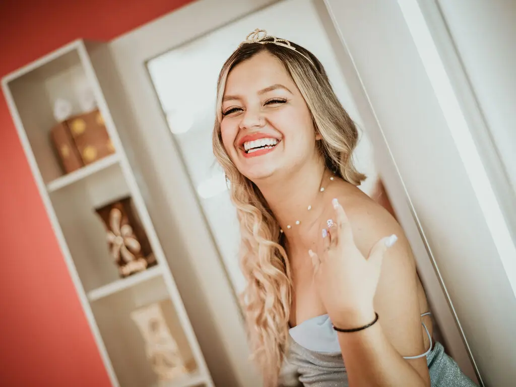 Smiling blonde woman in a silver dress laughing in a bright room with gift boxes.