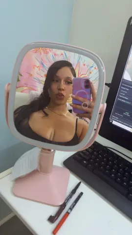 Young woman with long dark hair taking a selfie in a pink vanity mirror at her desk setup.