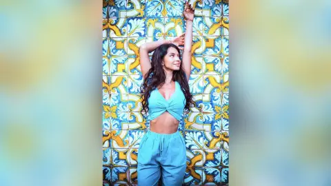 Cheerful woman in a teal crop top and lounge pants posing against vibrant blue and yellow patterned tiles.