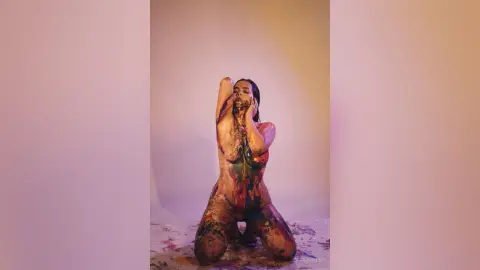 Artistic portrait of a woman covered in colorful paint posing against a soft pastel studio background.