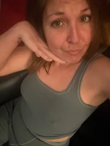 MistressBitchcoin wearing a grey tank top, resting her chin on her hand while looking at the camera.
