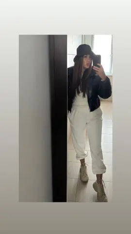 Woman in white loungewear, black puffer jacket, and bucket hat taking a mirror selfie in a modern room.