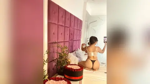 JessieOlivetti poses in black lingerie on a white bed, looking into a mirror in her bedroom.