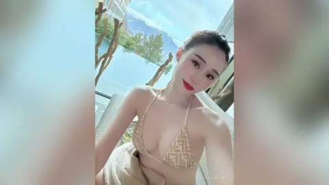 Young woman in a beige patterned bikini relaxing on a sunny balcony with a scenic mountain view.
