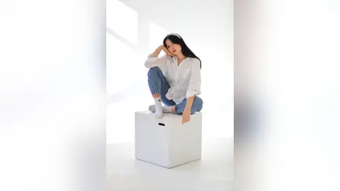 Young woman in white shirt and blue jeans sitting on a white cube in a bright, minimalist studio setting.