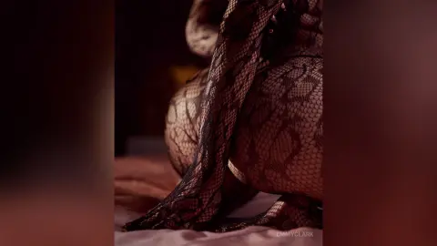 Woman in black lace patterned tights sitting on a bed in a moody, dimly lit bedroom setting.