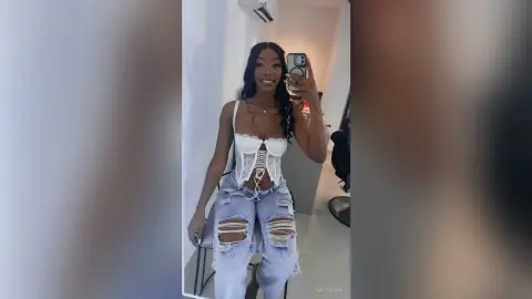 NatyGlow poses in a white lace corset and ripped blue jeans, showing off her long dark hair in a mirror selfie.