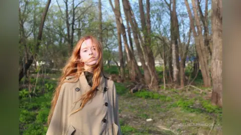 Redheaded woman in a beige trench coat standing in a sunny spring forest setting.