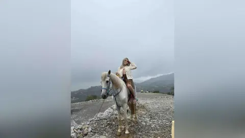 Blonde woman in a white jacket riding a white horse through a misty mountain landscape.