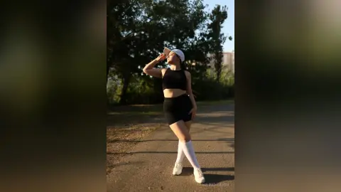Athletic woman in black crop top and shorts posing in a sunny park during golden hour.