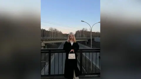 Blonde woman in a long black coat standing on a bridge at dusk against a soft twilight sky.