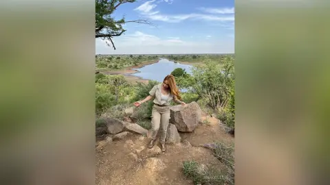 Cheerful woman in a beige jacket and trousers standing on a rocky cliff overlooking a scenic river valley.