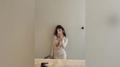 Young woman in a chic beige long-sleeve dress taking a stylish mirror selfie in a minimalist room.