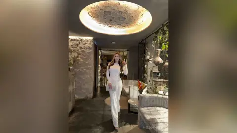 Elegant woman in a white jumpsuit standing in a modern luxury lounge with moon-themed ceiling lighting.