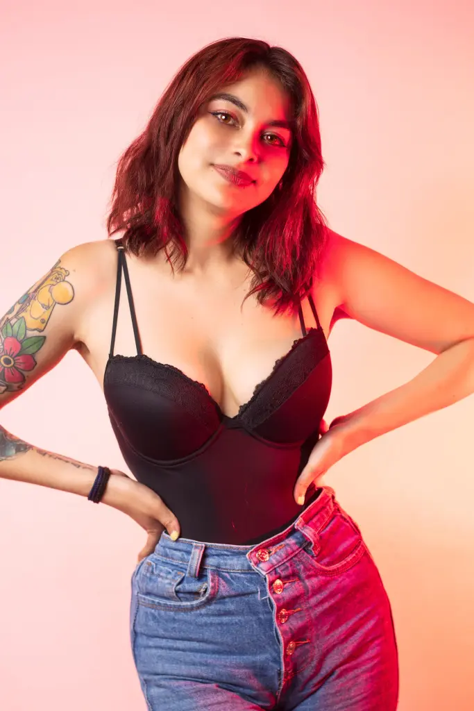 Glamorous woman in a black bodysuit posing against a warm pink studio background with moody lighting.