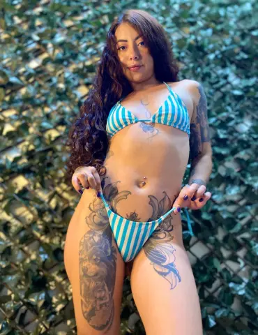 Tattooed woman in a blue striped bikini posing against a lush green leafy background.