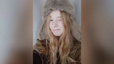 Smiling blonde woman in a cozy grey faux fur hat and winter coat posing for a warm portrait.