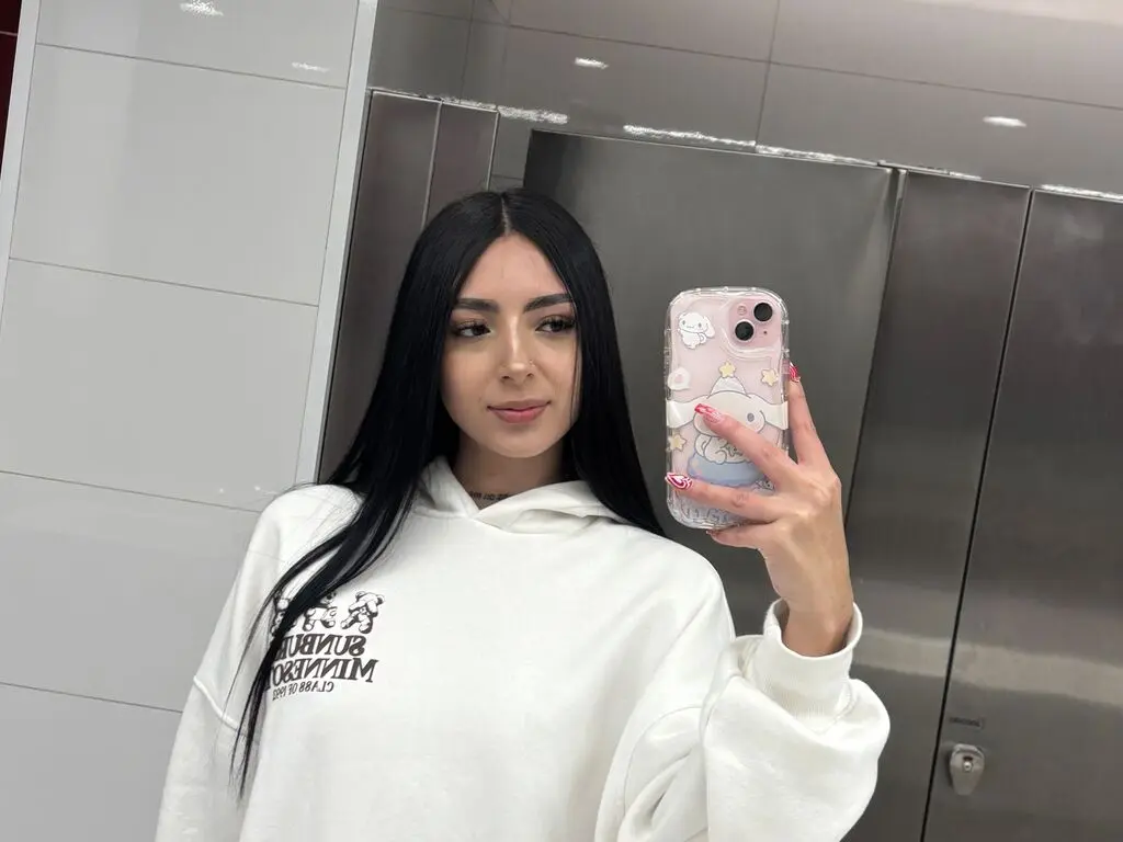 Young woman with long dark hair wearing a white graphic hoodie taking a mirror selfie in a modern elevator.